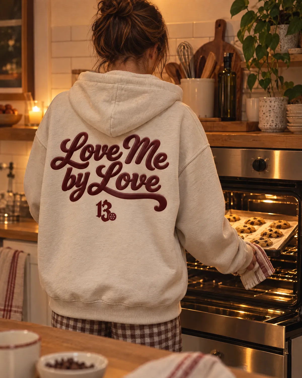 Seen from behind at a warm kitchen oven, sliding out a tray of cookies, in a bone hoodie with Love Me by Love in oxblood script on the back.